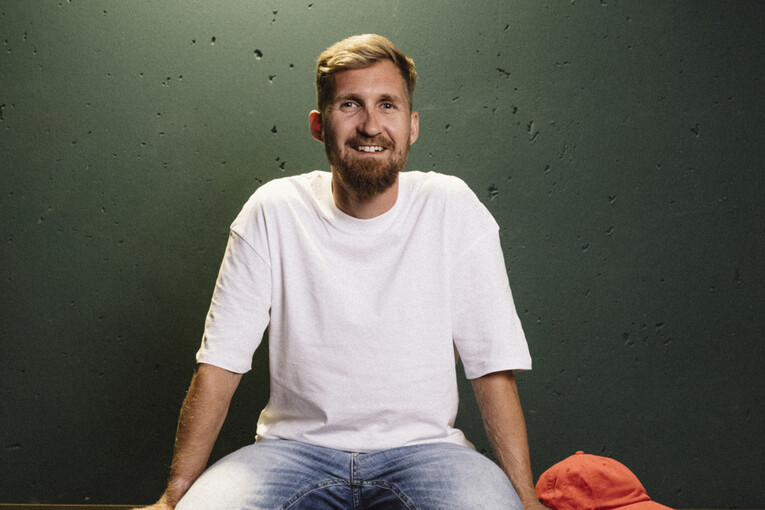 Senior Product Owner sitzt lässig auf einem dunkelgrünen Regal und lächelt in die Kamera