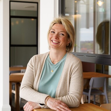 Bereichsvorstandsassistenz in Technologie steht vor einem Fenster. Sie stützt sich auf einem Tisch ab und lächelt in die Kamera