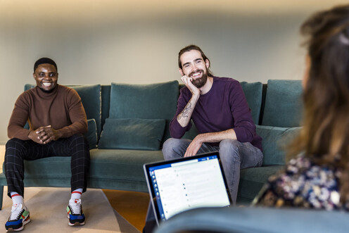 Zwei Kollegen sitzen auf einem Sofa und blicken zu ihrer Kollegin, die schräg gegenüber sitzt mit ihren Laptop
