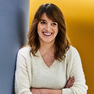 Unsere Kollegin, eine Quereinsteigerin, hat die Arme verschränkt, lehnt an der Wand und lacht zur Seite.