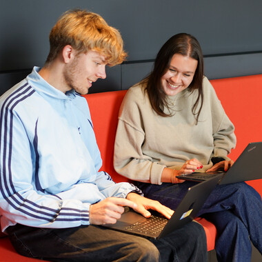 Zwei Werkstudenten eine Frau und ein Mann sitzen auf einem roten Sofa mit ihren Laptops und tauschen sich aus