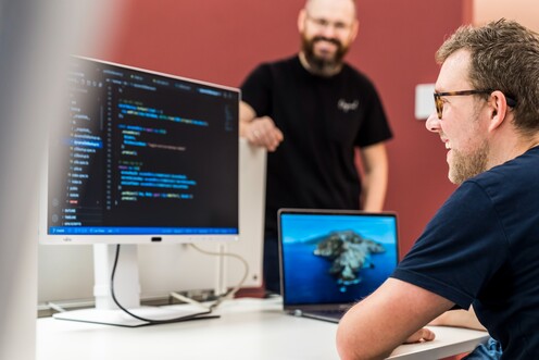 Ein IT-Mitarbeiter sitzt am Schreibtisch und guckt auf den Code, der auf dem Computerbildschirm angezeigt wird. Ein anderer Kollege steht freundlich daneben.