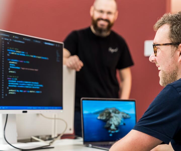 Software-Entwickler arbeiten als Team an der Weiterentwicklung unserer Plattform Ein IT-Mitarbeiter sitzt am Schreibtisch und guckt auf den Code, der auf dem Computerbildschirm angezeigt wird. Ein anderer Kollege steht freundlich daneben.