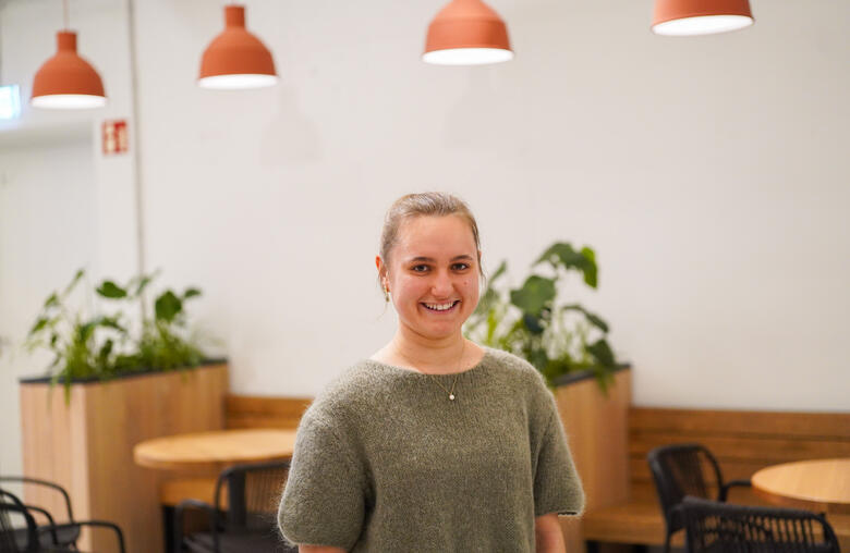 Eine duale Studentin der Business Administration steht im Social Space und lächelt in die Kamera. Im Hintergrund sind mehrere Holztische und Sitzmöglichkeiten, die mit Pflanzen dekoriert sind, zu sehen. 