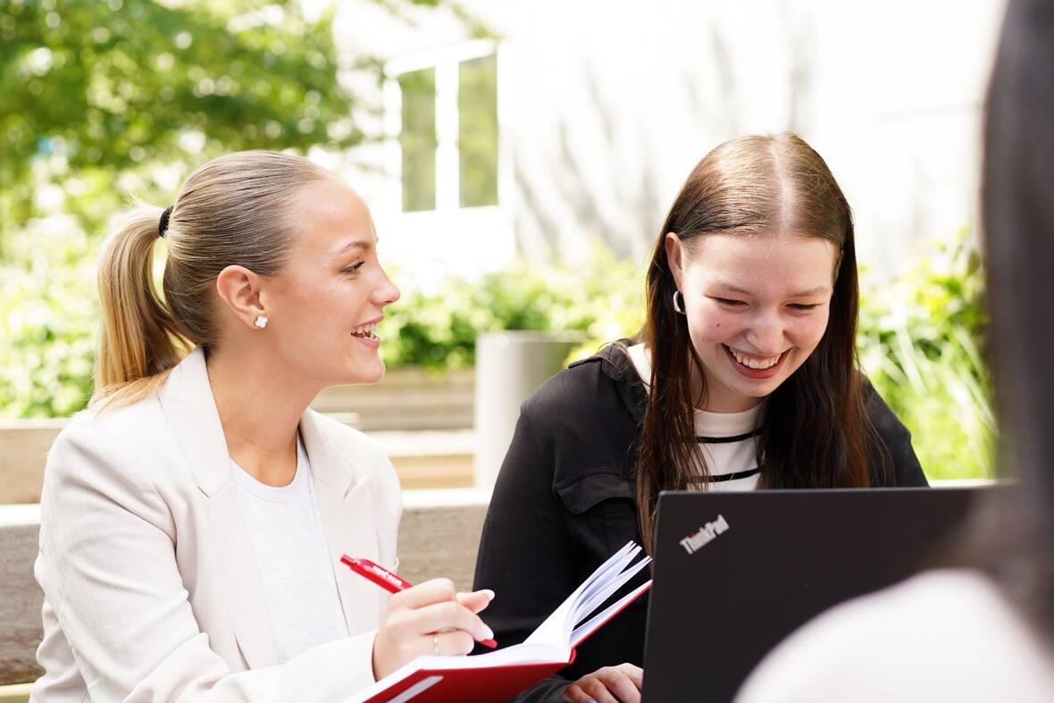 Das Bild zeigt zwei lachende Studentinnen auf einer Bank mit einem Notebook und einem Buch und Stift beim lernen auf dem Otto Campus. 