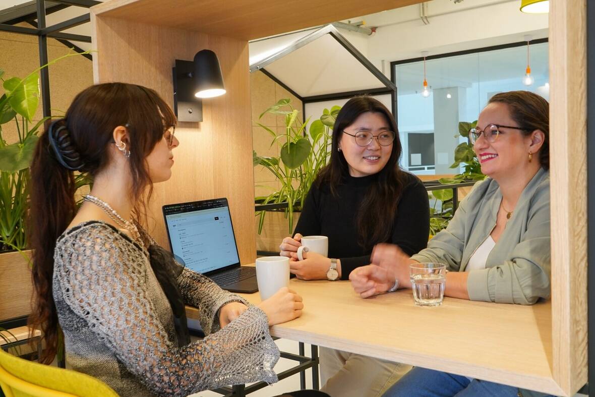 Drei Kolleginnen aus dem UX Design sitzen im Social Space an einem Tisch mit Laptop und Getränk und unterhalten sich. Im Hintergrund sind mehrere Pflanzen. 