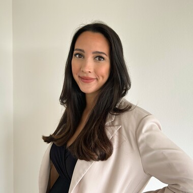 Unsere Kollegin als Process Lead IT-BI steht vor einer weißen Wand und hat deine Hand in der Hüfte und lächelt in die Kamera