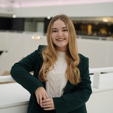 Unsere Kollegin ist Junior HR-Managerin im PM-Team. Sie lehnt sich mit dem Arm am Geländer ab und lächelt in die Kamera. Sie befindet sich im Headquarter Gebäude von OTTO.