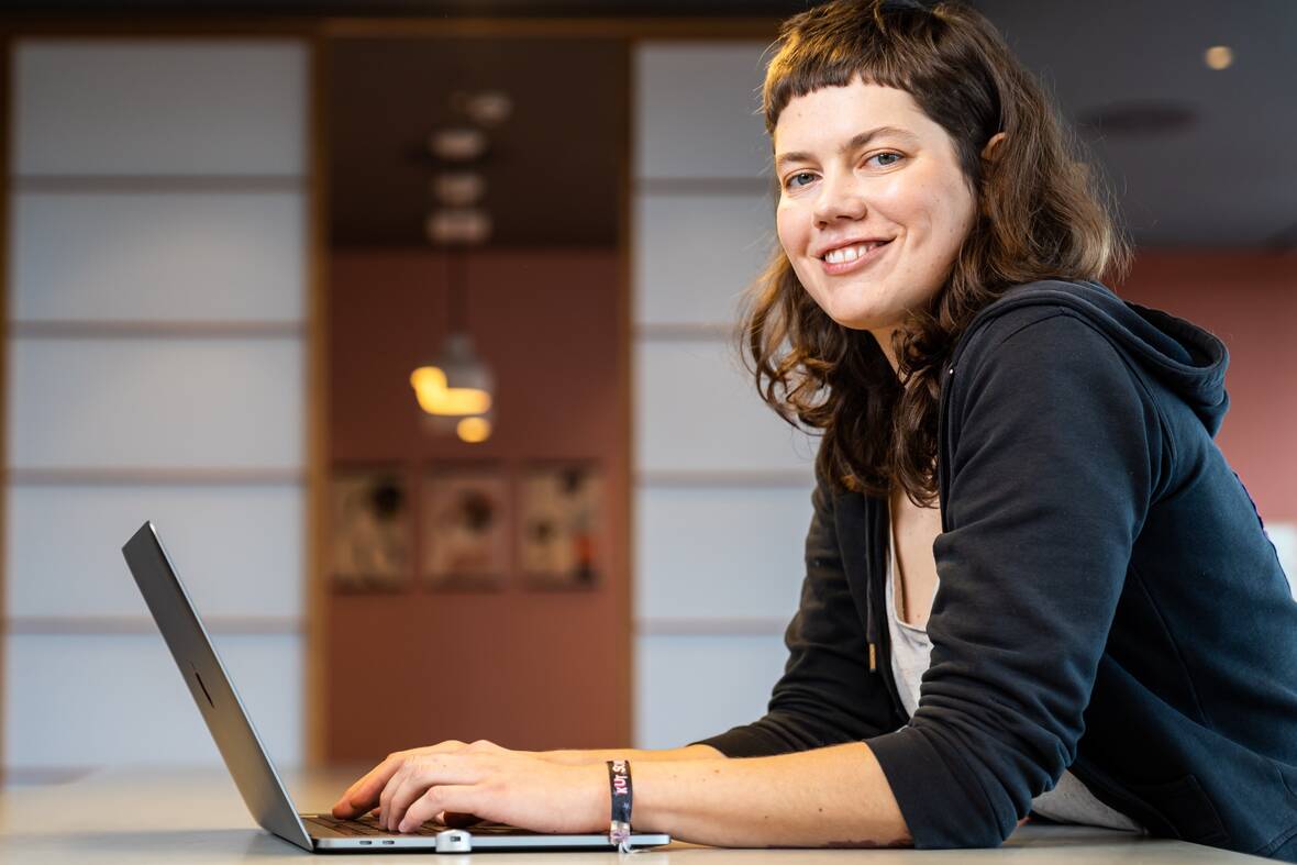 Softwareentwicklerin sitzt am Computer und guckt freundlich zur Seite in die Kamera.