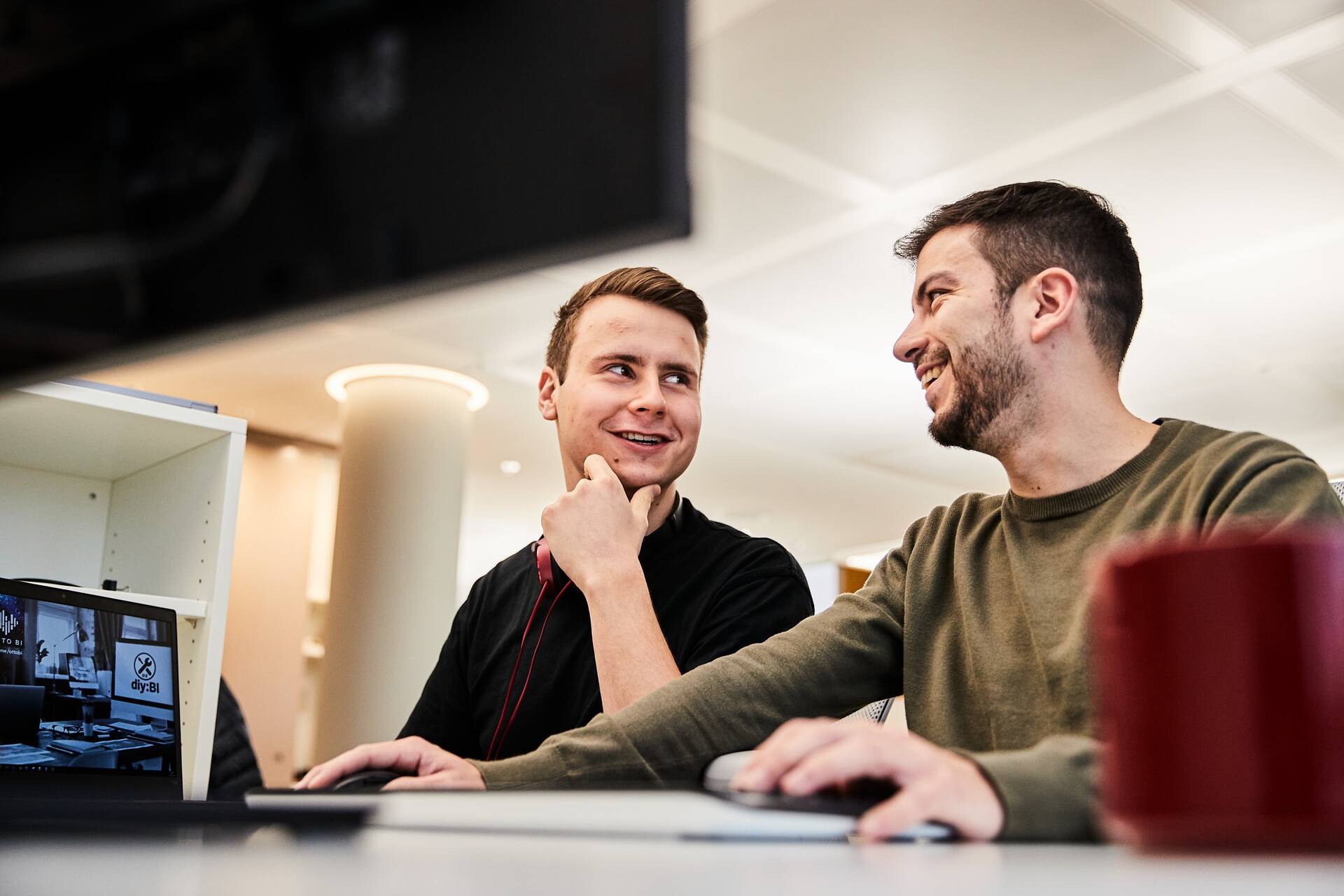 Zwei Kollegen aus Tech sitzen gemeinsam vor dem PC und unterhalten sich lächelnd.