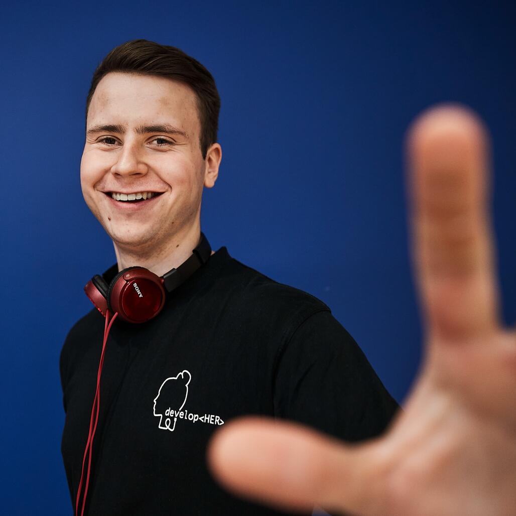 Unser Kollege in Tech Unser Kollege steht fröhlich vor einem blauen Hintergrund und streckt den Arm nach vorne.