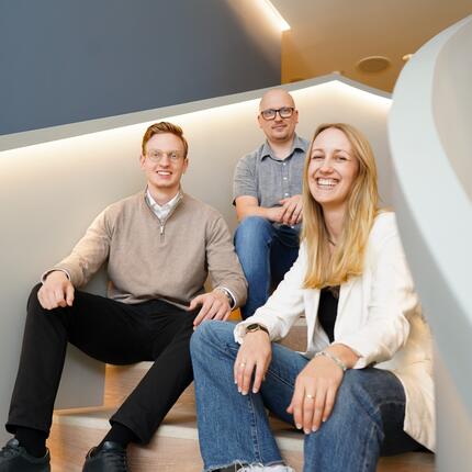 Drei Kollegen sitzen auf der Treppe und lächeln in die Kamera.