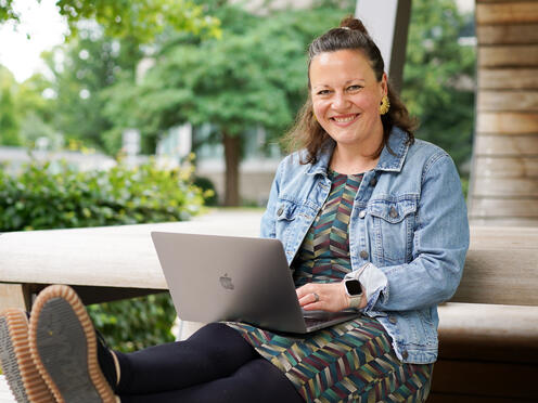 Eine Expertin Technical sitzt  lächelnd mit ihrem Laptop auf einer Holzbank auf dem Otto Campus.