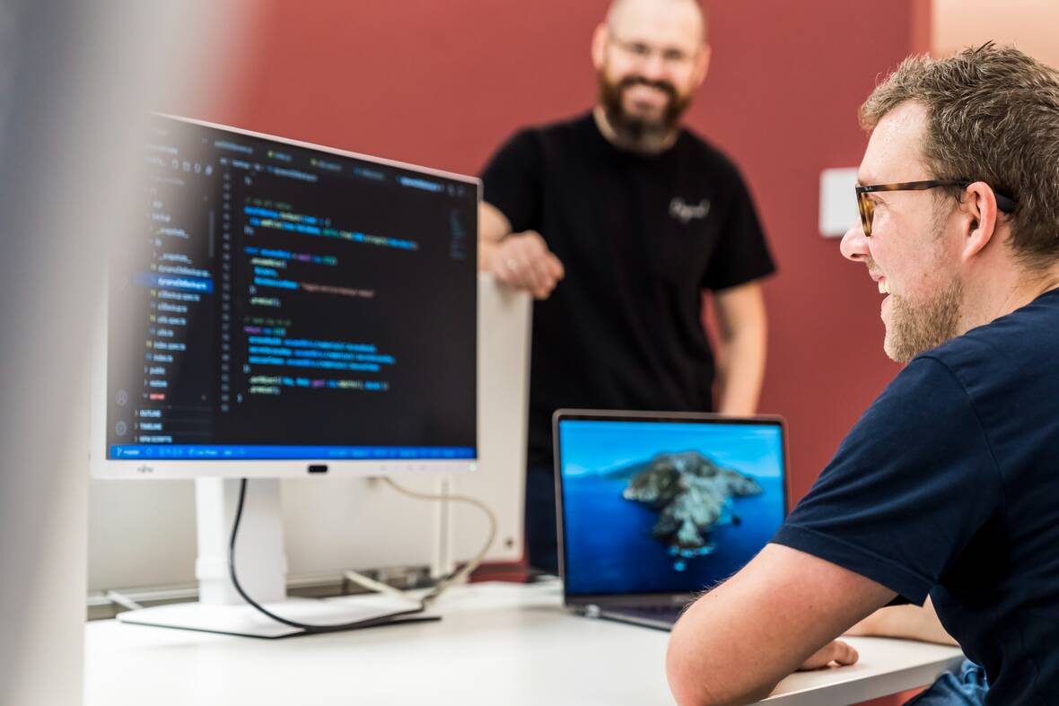 Ein IT-Mitarbeiter sitzt am Schreibtisch und guckt auf den Code, der auf dem Computerbildschirm angezeigt wird. Ein anderer Kollege steht freundlich daneben.