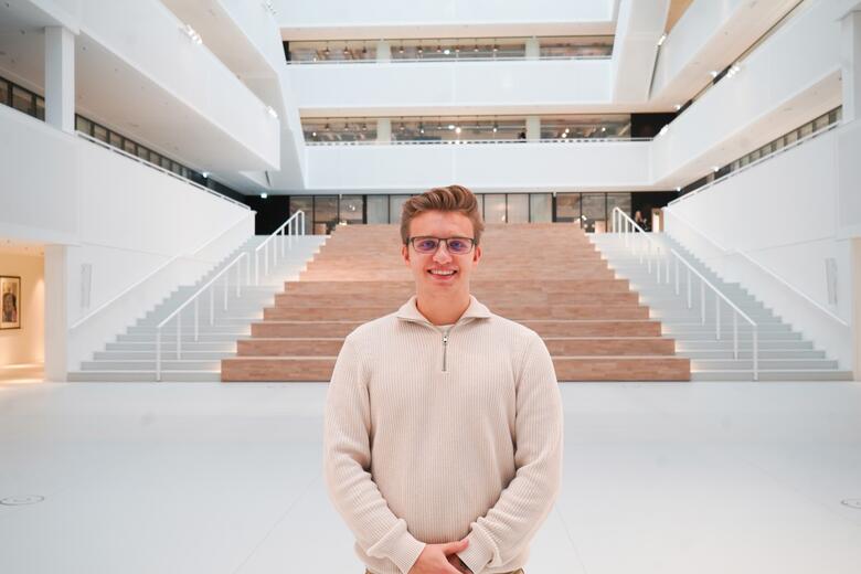Junior Software Developer steht mittig im Atrium im neune Gebäude 4 unser Headquarter Gebäude von OTTO und lächelt in die Kamera.
