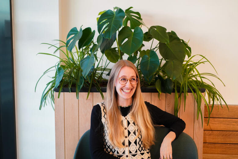 Duale Studentin aus Business Administration sitzt von einem Stuhl vor einer Holzwand mit Pflanzen und lächelt zur Seite