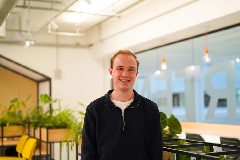 Ein dualer Student der Fachrichtung Business Administration bei OTTO lächelt in die Kamera. Im Hintergrund sind Pflanzen und verschiedene Lichter zu sehen. 