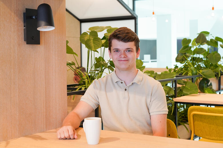 Dualer Student der Wirtschaftsinformatik sitzt lächelnd mit seiner Tasse in einem unserer Social-Space-Räume, in dem im Hintergrund mehrere Pflanzen zu sehen sind.