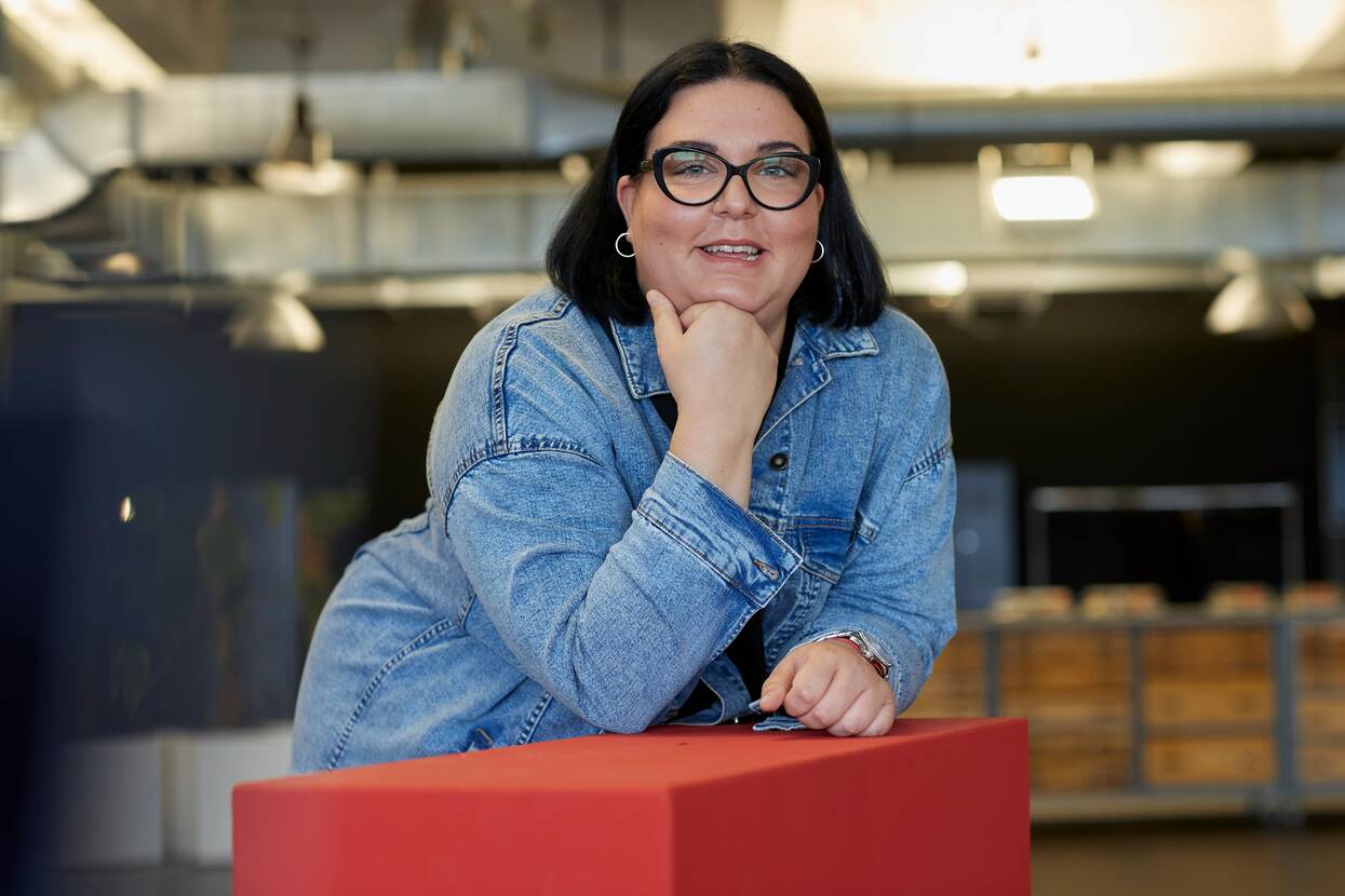 Unsere Kollegin arbeitet im Callcenter von OTTO, trägt eine Brille und lehnt lächelnd auf einem roten Hochtisch.