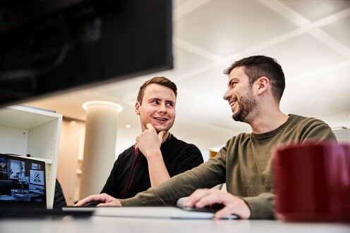 Unsere Kollegen in Tech beim Arbeiten Zwei Kollegen aus Tech sitzen gemeinsam vor dem PC und unterhalten sich lächelnd.